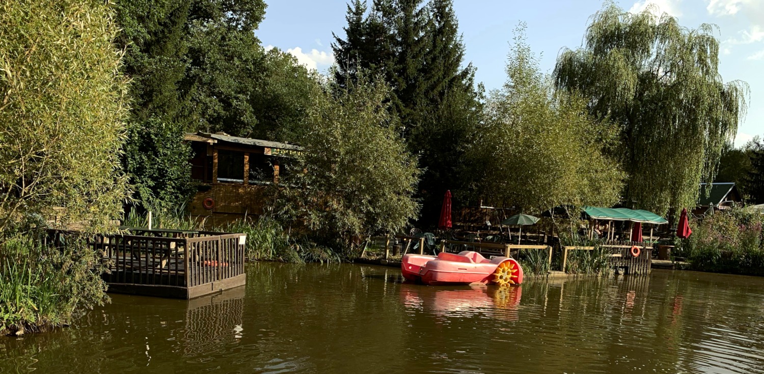 Teich im Wald: Anlegestelle am Ufer mit verschiedenen Holz-Booten. In der Mitte ein rotes Tretboot.