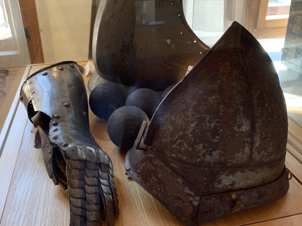 Ritterrstung, bestehend aus Handschuh, Brustplatte (hinten) und Helm (vorn). Dazwischen liegen Kanonenkugeln aus Metall.