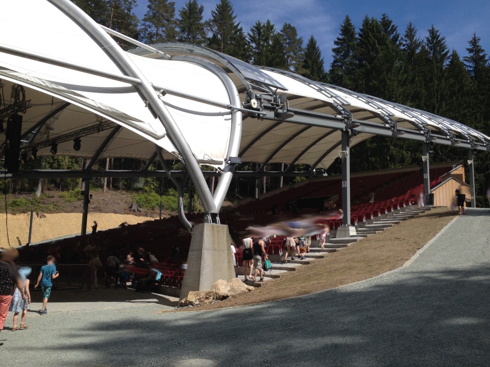 Stahlkonstruktion mit weißem Dach auf schräger Bühne hinter Ascheplatz. Im Hintergrund Wald.