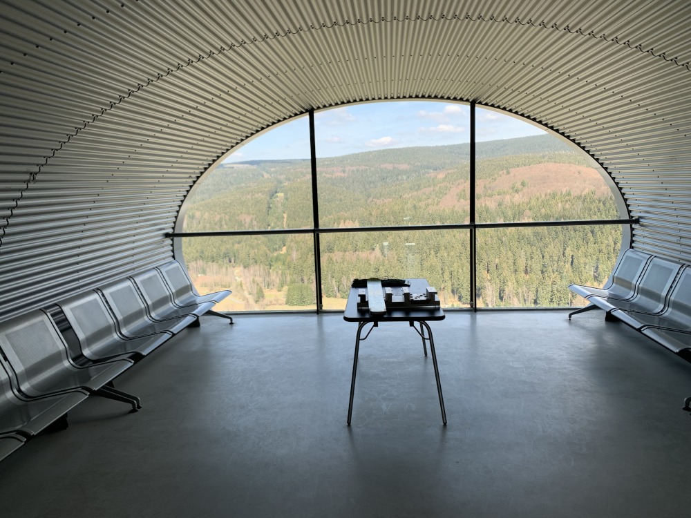 Rhrenfrmiger Aufenthaltsraum aus Metall mit Metallsitzen an den Seiten. Am Ende eine ovale Fensterfront mit Ausblick auf den Wald.