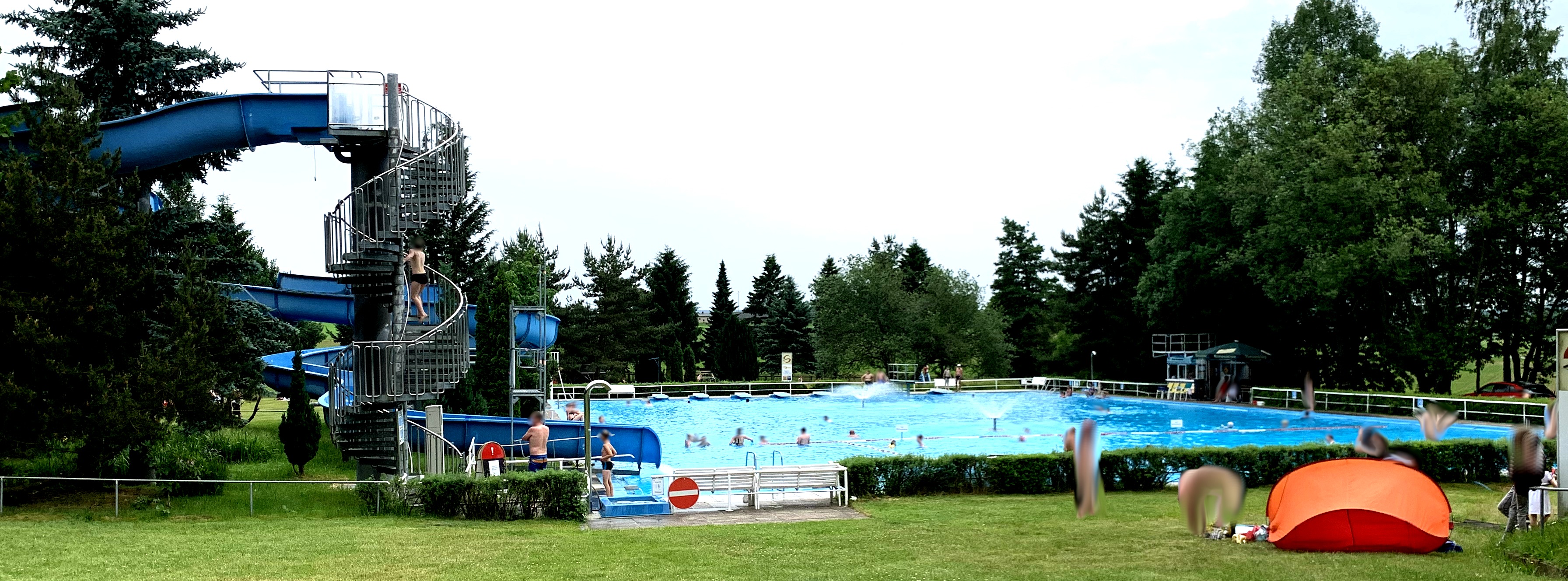 Schwimmbecken inmitten von Wiese und Bäumen. Links Wendeltreppe mit Rutsche