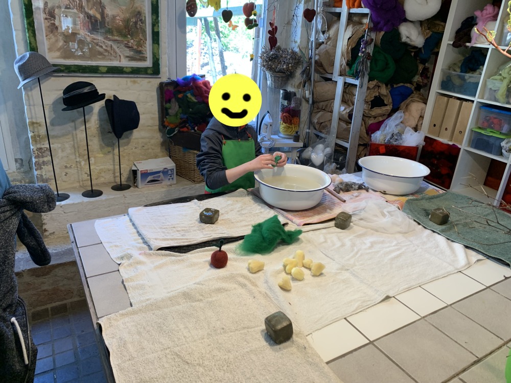 In der Werkstatt bastelt ein Junge ber einer Schssel mit Wasser. Auf dem Tisch liegen die Materialien Filz und Seife verteilt. Das Gesicht des bastelnden Kindes ist mit einem Smily unkenntlich gemacht. Im Hintergrund Hte und Bastelmaterialen im Regal.