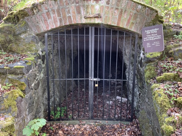 Eingang einer Höhle, von oben mit Ziegel gestützt, verschlossen von einem schwarzen Tor mit Schloss