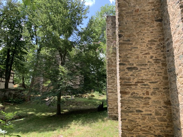 Links: durch Bäume Ruine durchschimmernd - rechts hohe Steinmauer bis nach oben