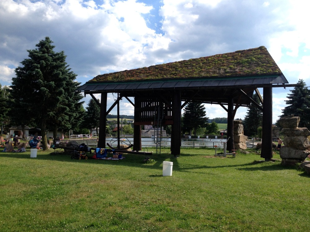 Wiese, Kletterhaus mit einer Ebene, Baum, Himmel. - Im Hintergrund ein Schwimmbecken.