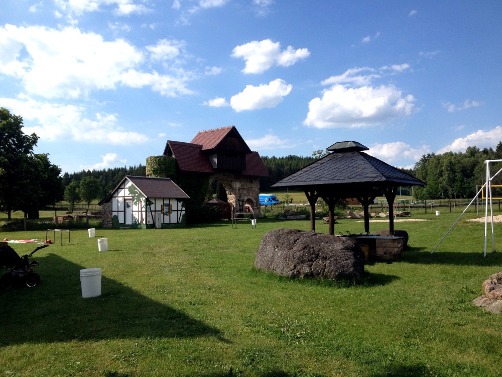 Wiese. Darauf ein groer Stein, berdachter Sitzbereich, ein Gebude. Im Hintergrund Wald. Blauer Himmel mit Wolken.