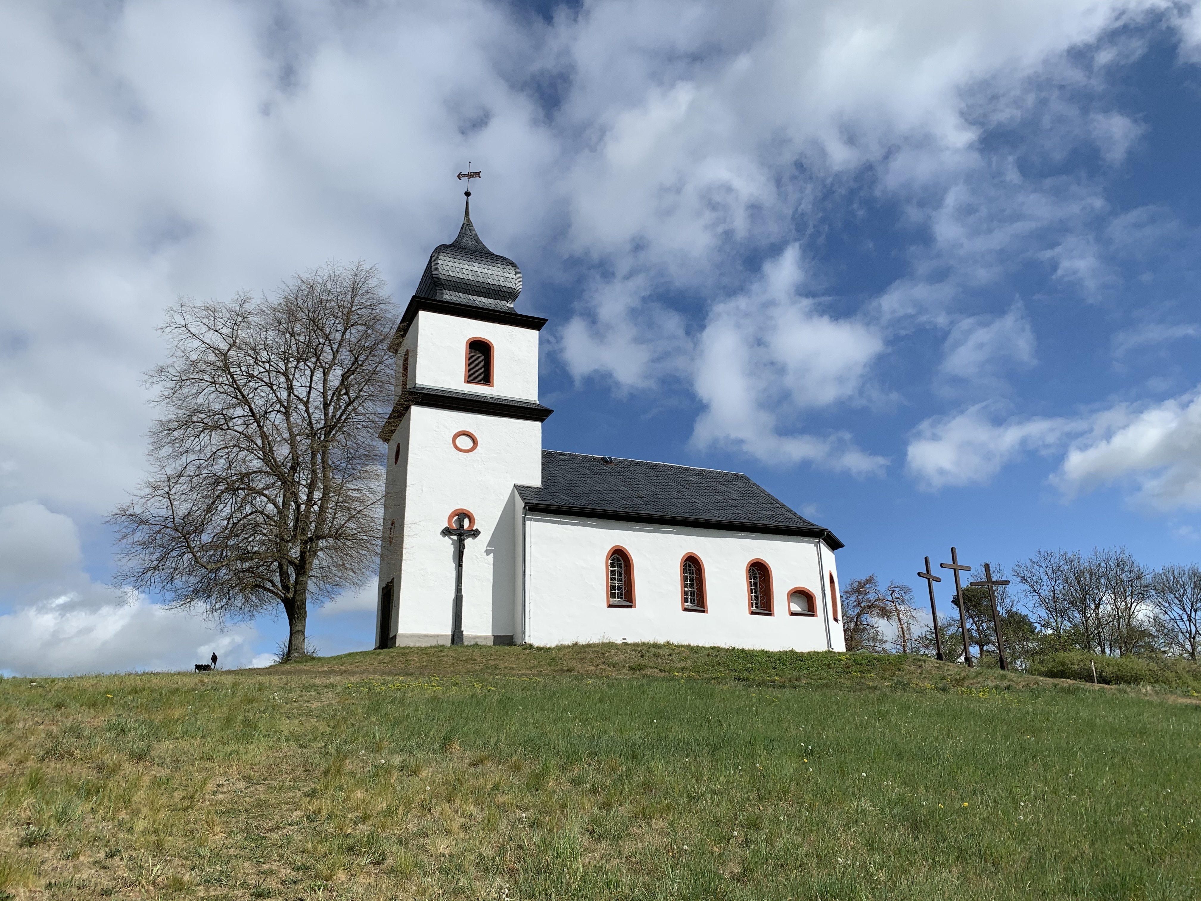 Kapelle auf grasbewachsenem Hügel unter Himmel. Kirchenhaus zwischen einzelnen Bäumen und 3 Kreuzen.