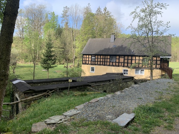 Weg, Wiese, Bume und Wohnhaus. In der Mitte befindet sich das Dach einer steinernen Konstuktion.