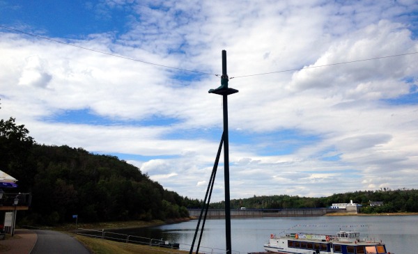 Unter bewlktem Himmel Holzstange mit Platform, welche mit 2 Seilen verbunden ist. Unten links ist ein Weg zu sehen und rechts unten ist ein Schiff auf dem Wasser.