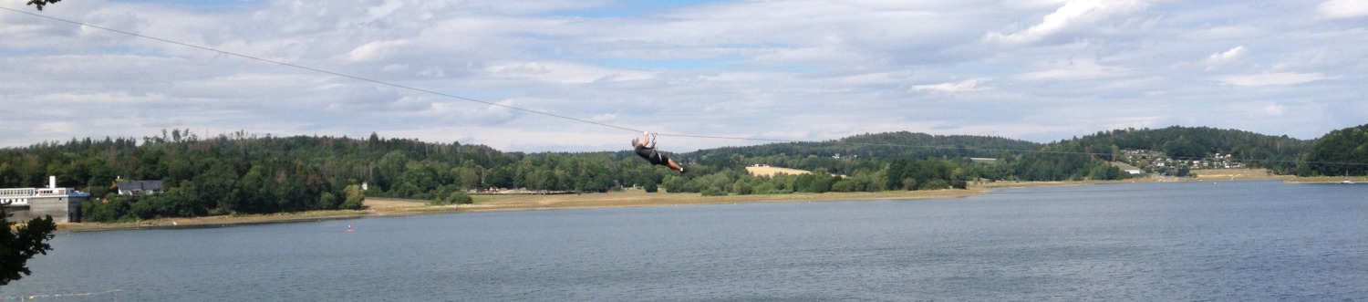 Sportler rutsch gesichert an einem Seil von links nach rechts. Im Hintergrund sind Wolken, Wald und Wasser zu sehen.