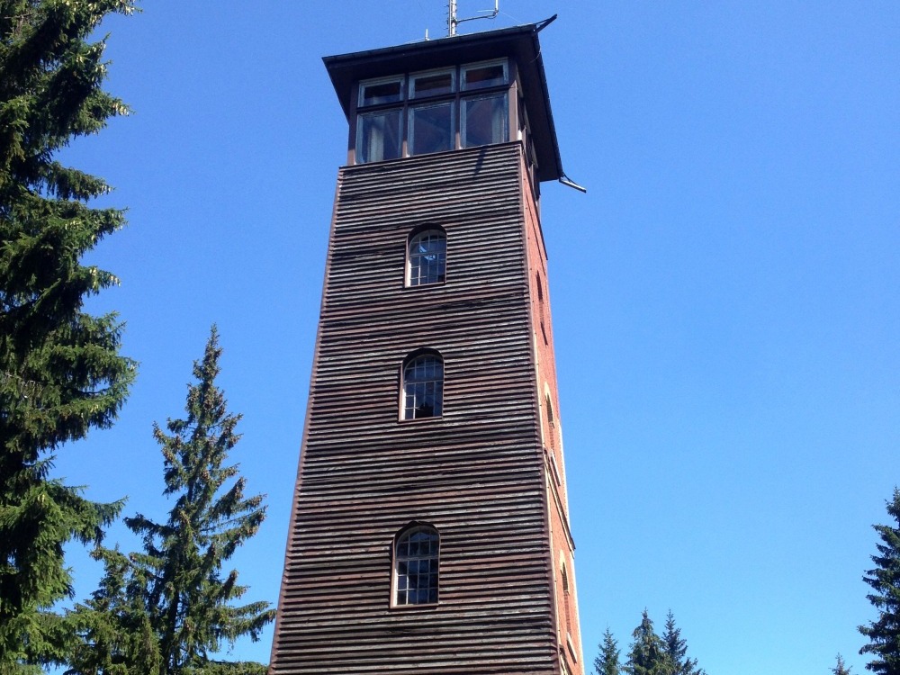 Turm hinter Teil einer Hütte. Auf dem Turm befindet sich eine Sende-Antenne. Links und rechts stehen Bäume.