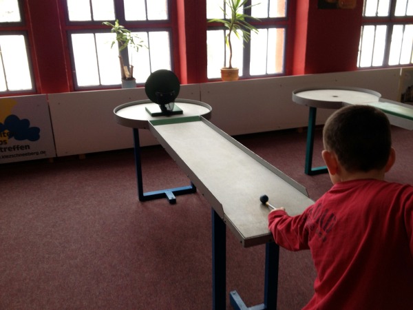 Junge in rotem Pullover spielt Kugel auf Pitpat-Tisch in Richtung Sprungrampe. Im Hintergrund sind Fenster.