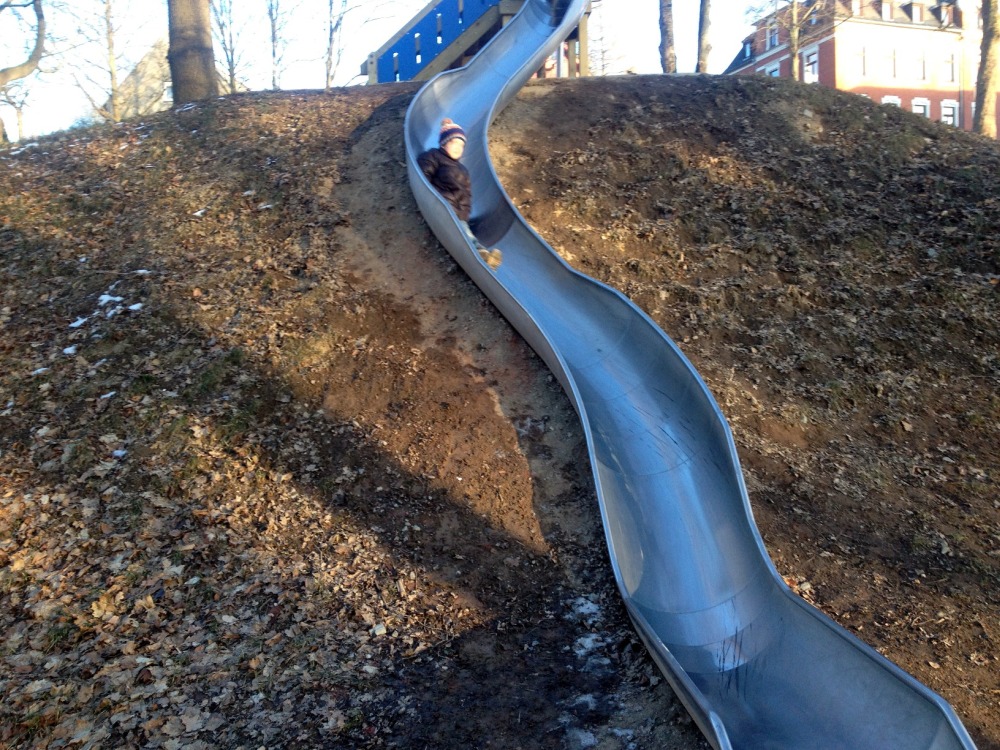 Kind rutscht auf langer Metall-Rutsche den Berg hinunter.