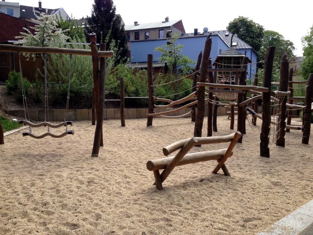 Schaukel und Klettergerst, davor Sitzbank. Alles aus Holz. Weicher Sand als Untergrund.