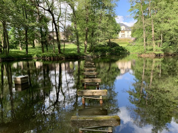 Balken stegartig ber einem Teich. Im Hintergrund Wiese und Herrenhaus.