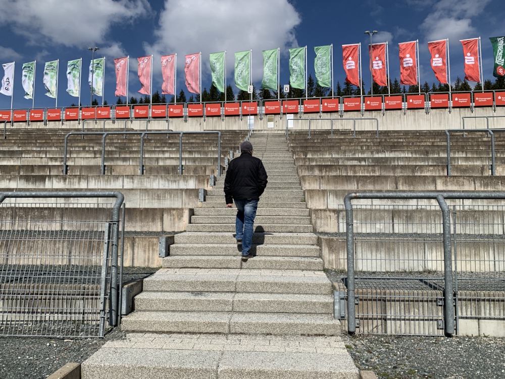 Mann luft auf Treppe nach oben. Links und rechts leere Besuchersitzpltze. Oben Flaggen von verschiedenen Sponsoren.