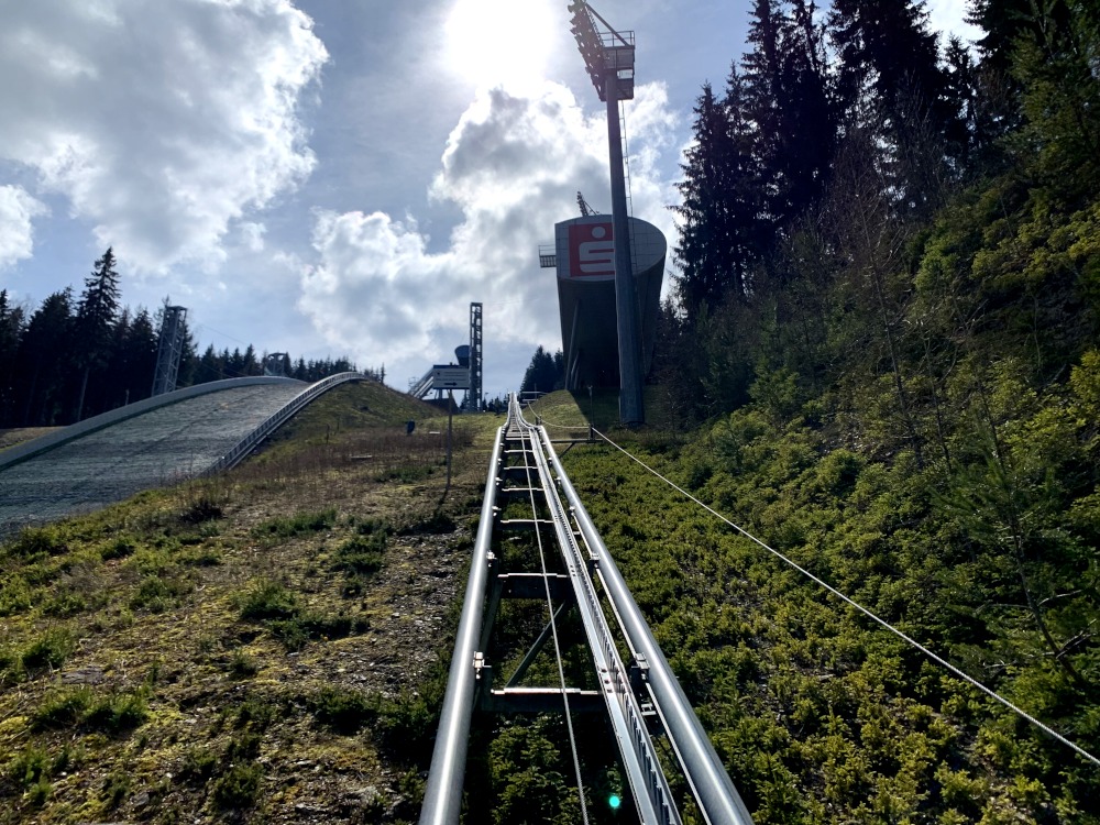 Stahl-Personenaufzug im Freien zwischen Schanzen-Hang und Wald. Rechts am Aufzug steht Wertungs-Kapsel und Lichtmast.