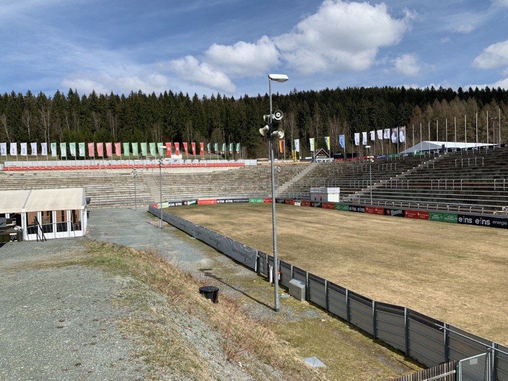 Auslauf einer Skisprungschanze. Darum leere Zuschauer-Tribne umrandet von Flaggen. Dahinter Wald.