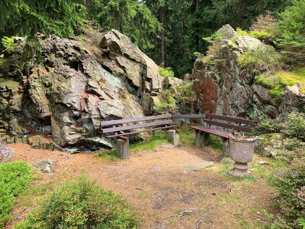 Bnke mit Mlleimer aus Stein vor Felsen vor Wald.